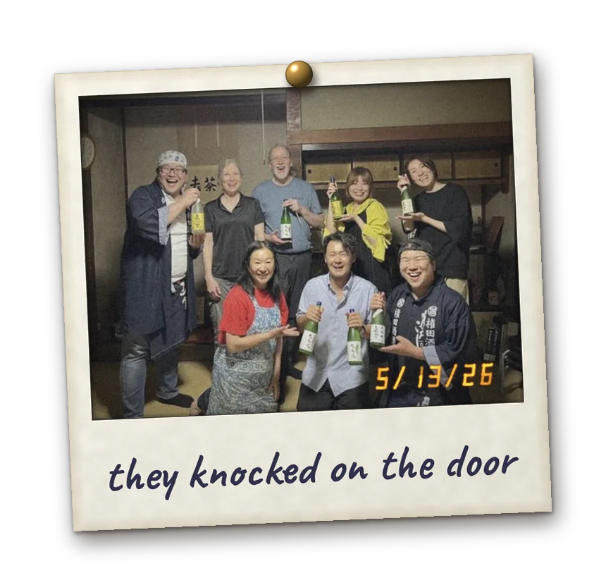 Polaroid pinned to the page with a brass tack: the Gonda family, our team and two guests from the U.S. hold bottles of Yumiyamaru in a tatami room. Date stamp 5/13/26. Handwritten: they knocked on the door